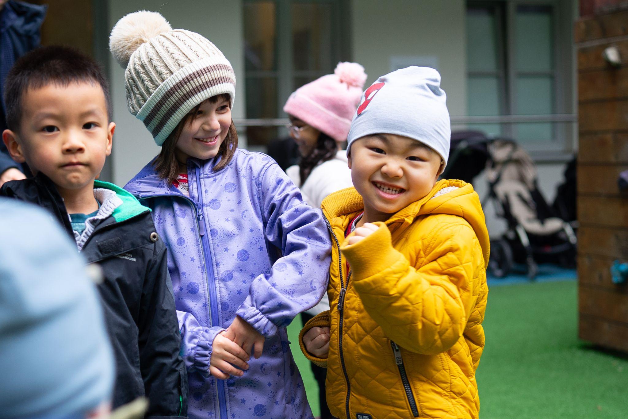 Drei Kinder in Winterjacken stehen an der Hand haltend im Hof vor einem Gebäude
